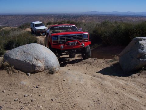 1985 Ford Bronco - Two off-road vehicles navigate a rocky trail with a scenic vista in the background.
