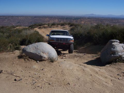 1985 Ford Bronco - White Ford Bronco navigates a rough off-road trail with large rocks and dry brush.