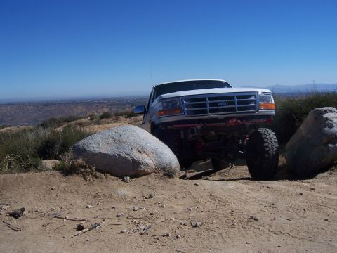 1985 Ford Bronco - A white Ford truck navigates a rocky, off-road trail under a clear blue sky.