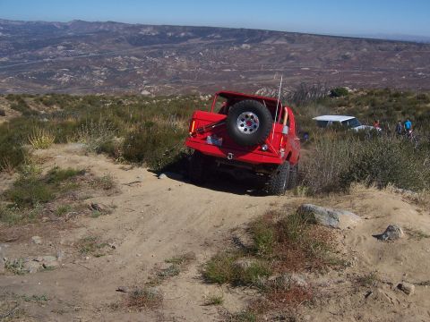 1985 Ford Bronco - A red SUV navigates a steep, dusty trail with a scenic mountain landscape in the distance.