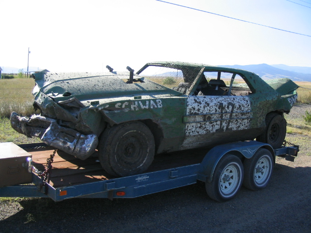 Green demolition derby car with crushed front end chained on a blue flatbed trailer