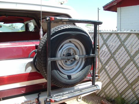 1990 Ford Bronco - Close-up of a black square-tube swing-away tire carrier bolted to the rear bumper of a maroon and white SUV, holding two spare tires on pla…