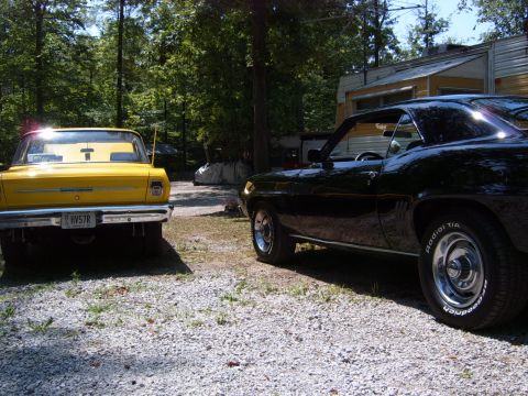 1993 Ford Bronco - A black 1969 Chevrolet Camaro is parked next to a yellow vehicle in a gravel lot with trees and a trailer in the background.