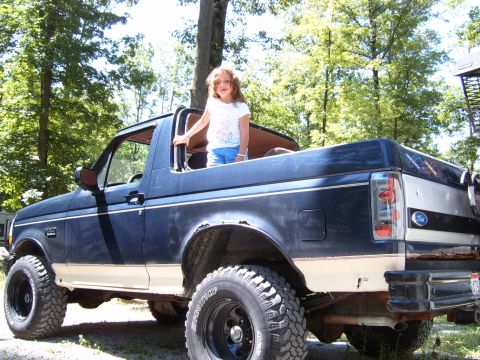 1993 Ford Bronco - A young girl stands in the bed of a dark blue and tan Ford Bronco, which is parked outdoors.