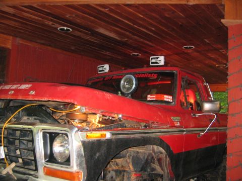 1979 Ford Bronco - A red Ford Bronco parked under a wood-ceilinged carport with its hood propped open over the engine bay, wearing a chrome grille surround, r…