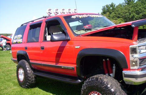 2006 Ford Supercrewzer - This red SUV, likely a Chevrolet Tahoe from the 1990s, features a lifted suspension, large off-road tires, and auxiliary lights on the roof.