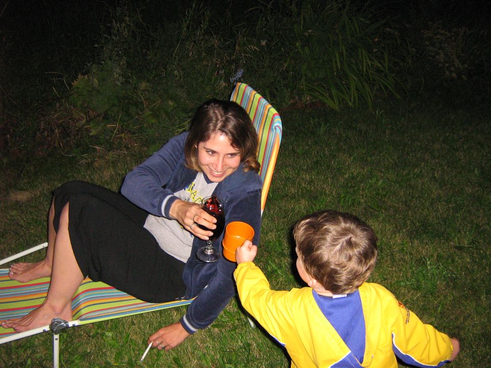 Woman sitting in a striped chair, holding a wine glass. A child in a yellow shirt offers an orange cup
