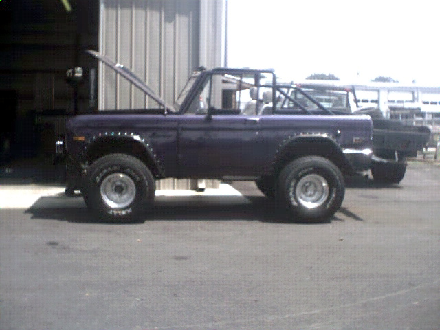 Purple Ford Bronco with hood open, roll cage, and aftermarket wheels. Another vehicle is parked behind it