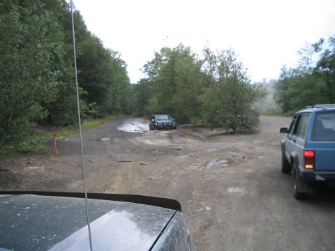 1992 Ford Bronco - Two SUVs navigate a muddy off-road trail surrounded by dense forest.