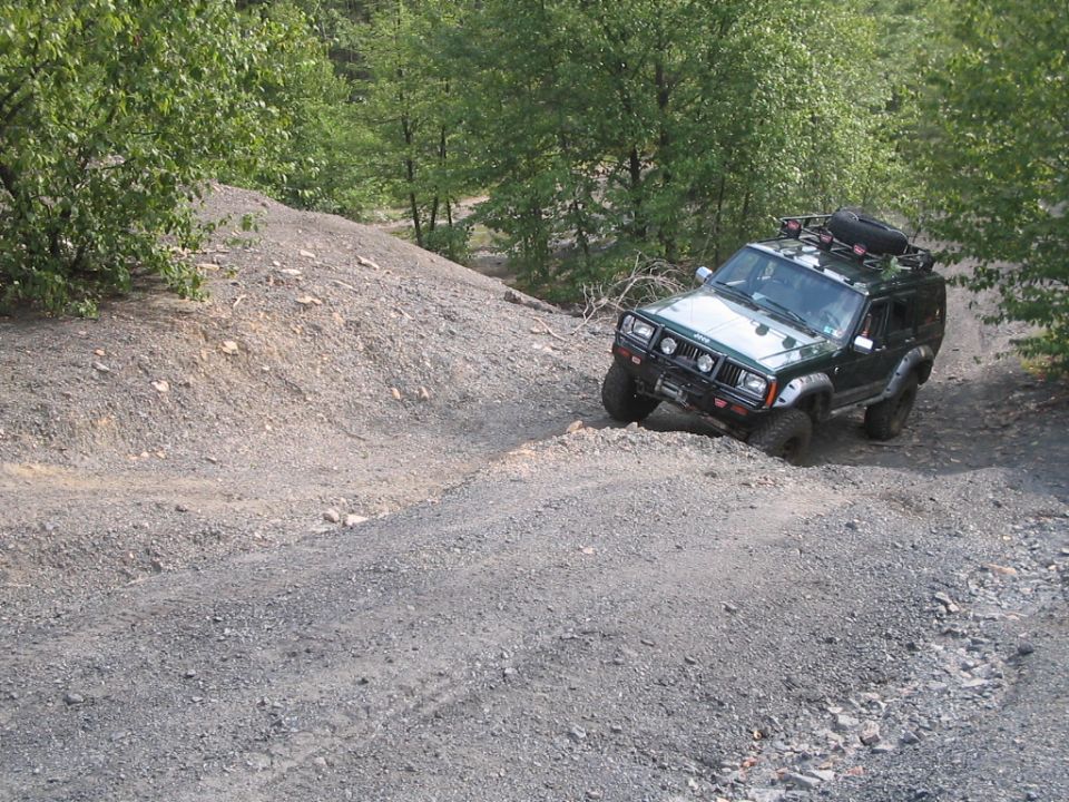 Dark green Jeep Cherokee XJ with roof rack and winch bumper descending a gravel hill off-road