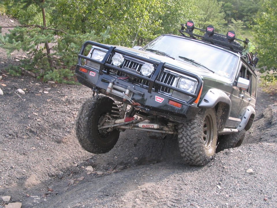 Green Jeep Cherokee XJ with winch bumper and light bar flexing on a rocky trail