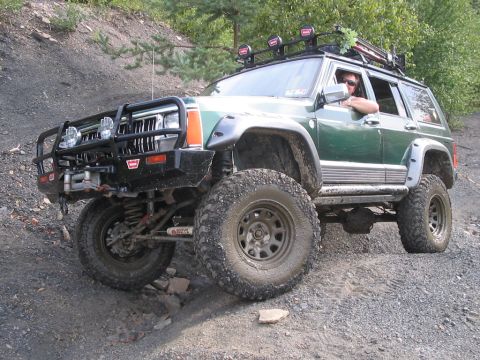 1992 Ford Bronco - A modified green Jeep Cherokee XJ navigates an off-road trail with large tires and a roof rack.