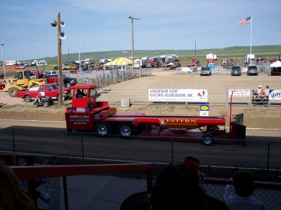 Red tow truck with a long trailer, parked at an outdoor event with other vehicles and tents in the background