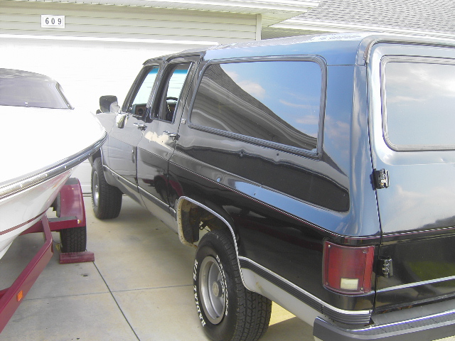 1990 Chevrolet Suburban parked next to a boat on a trailer, rear quarter view