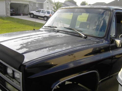 1990 Chevrolet Suburban - A black 1990 Chevrolet Suburban shows significant paint degradation on its hood, with another white SUV visible in the driveway.