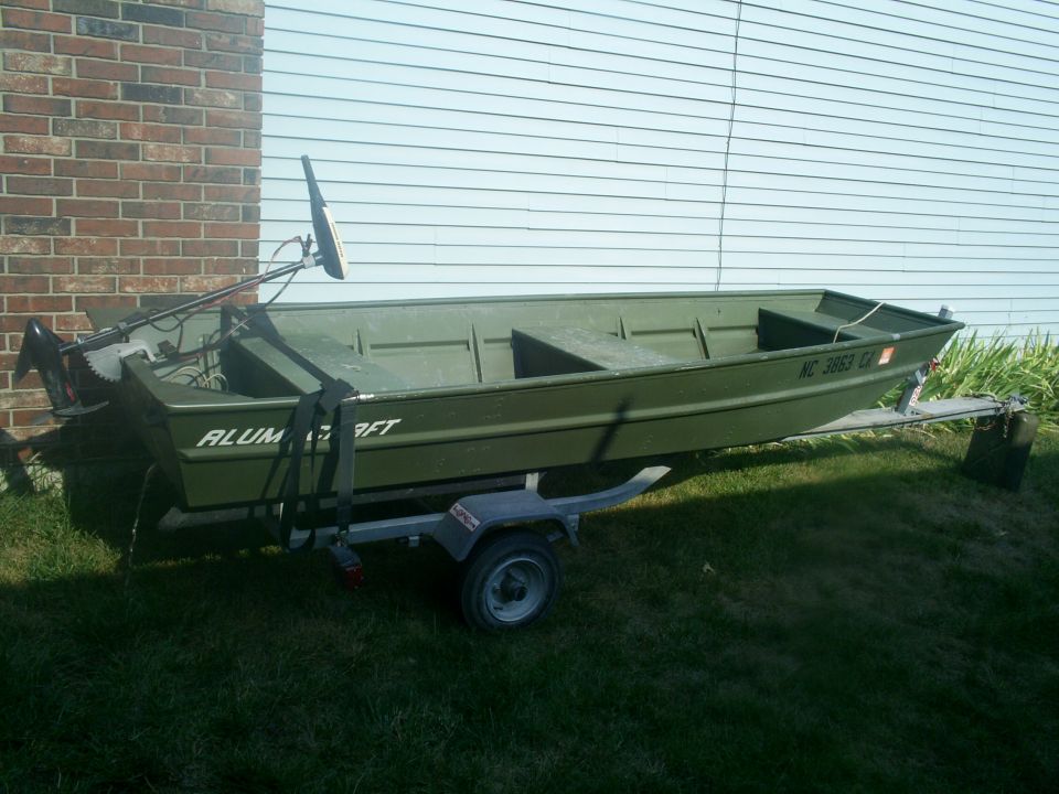 Olive green Alumacraft jon boat on a single-axle trailer by brick wall