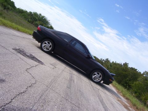 1994 Ford Probe - A black coupe with distinctive aftermarket wheels is seen from a three-quarter front angle on a broken asphalt surface.