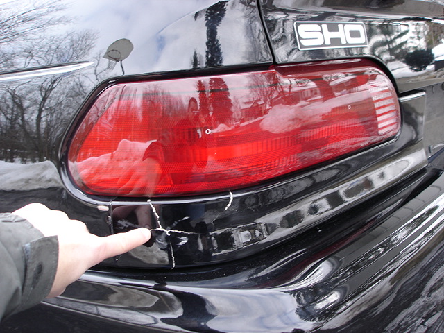 Close-up of cracked black rear bumper with a finger pointing to damage. SHO badge visible on car
