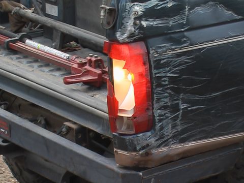 1981 Ford Bronco - A red hi-lift jack rests in the bed of a dark-colored truck next to a broken taillight and heavily scratched body panel.