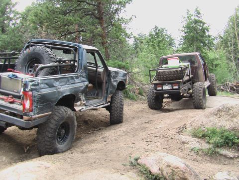1981 Ford Bronco - Two heavily modified off-road vehicles, including a 1981 Ford Bronco, navigate a rocky trail in a wooded area.