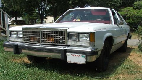 1985 Ford Crown Victoria LTD - This white Ford sedan, likely from the early 1980s, is parked on a grassy area with trees in the background.