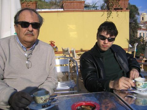 1969 Ford Mustang - Two men enjoy coffee at an outdoor cafe, with a yellow wall and potted plants in the background.