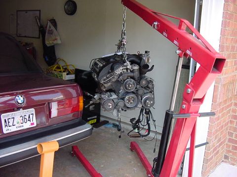 1984 BMW 3 Series - The rear of a maroon 1984 BMW 3 Series is visible next to a red engine hoist lifting its engine.