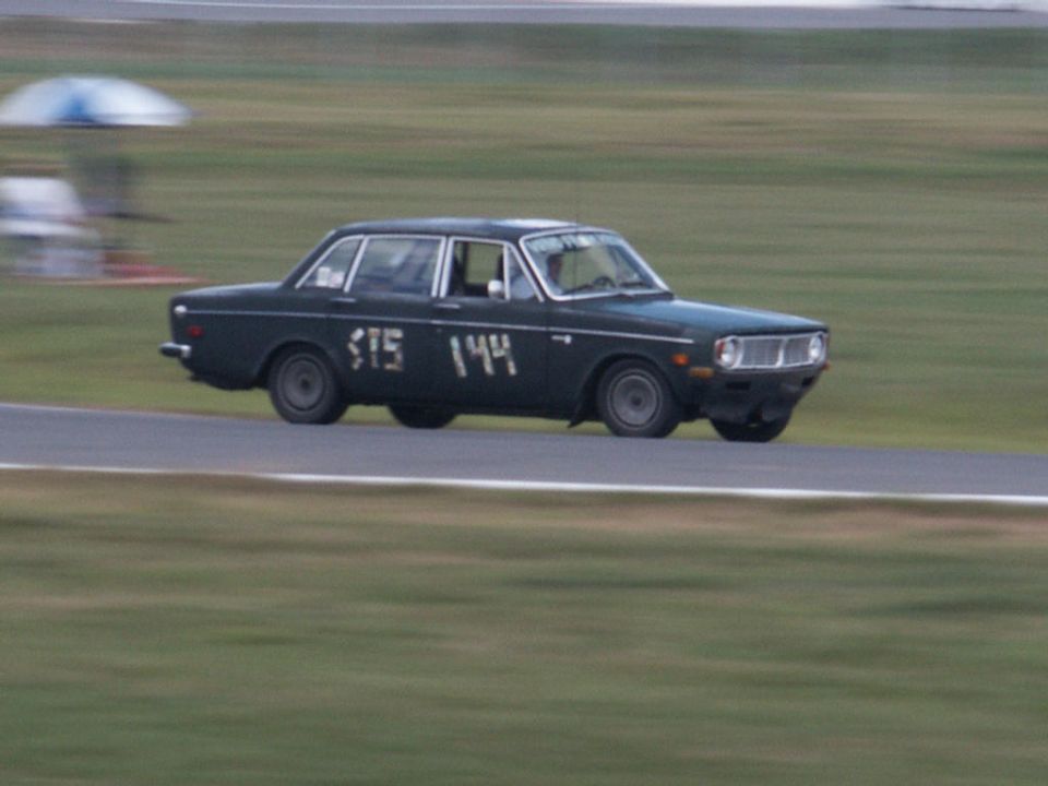 Dark green Volvo 144 sedan race car with painted numbers on a road course