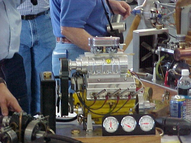 Scale model V8 engine with blower on a stand surrounded by people at a show