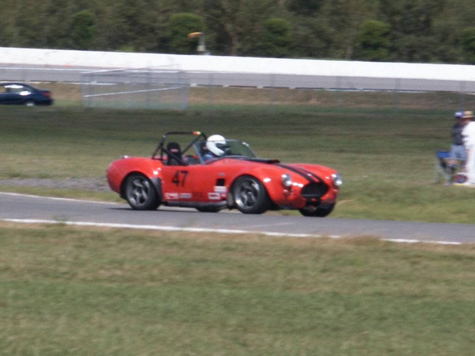 Red convertible race car with black stripes and number 47 on the side, driving on a track. Another dark car is visible in the background