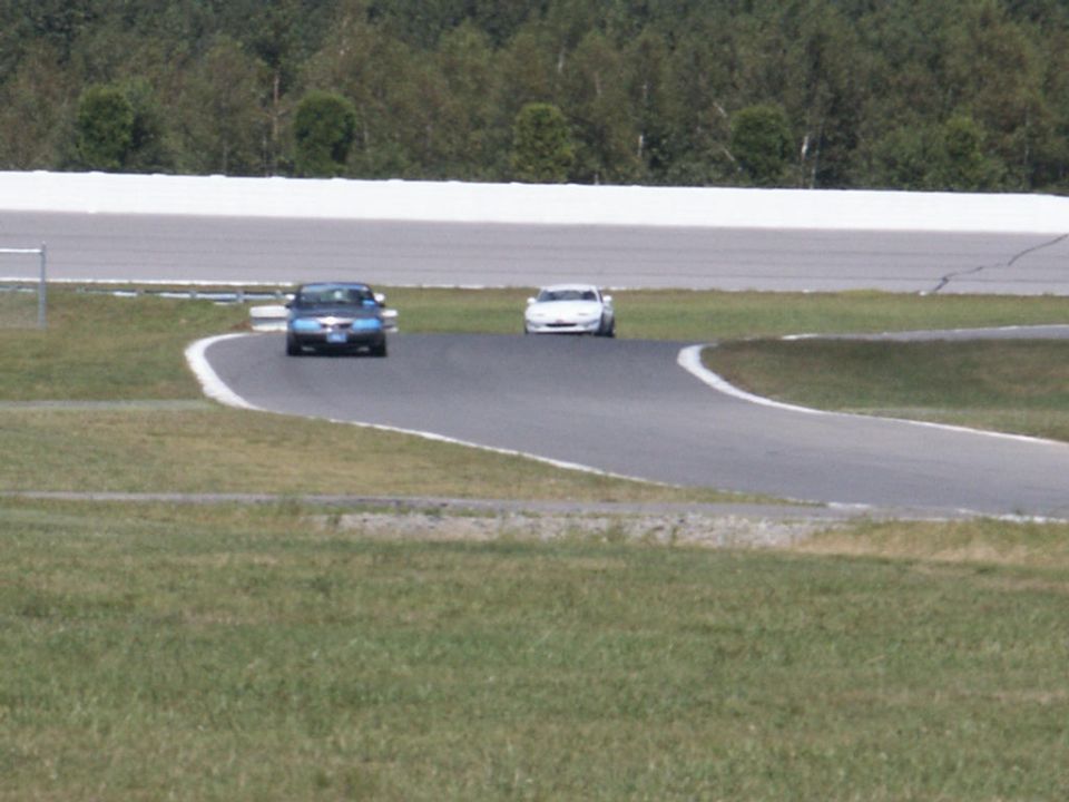 Blue sedan and white sports car on a racetrack, trees and white barrier in background