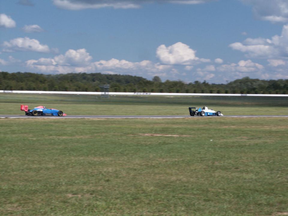 Two open-wheel race cars on a track, one blue with red accents, the other white and blue