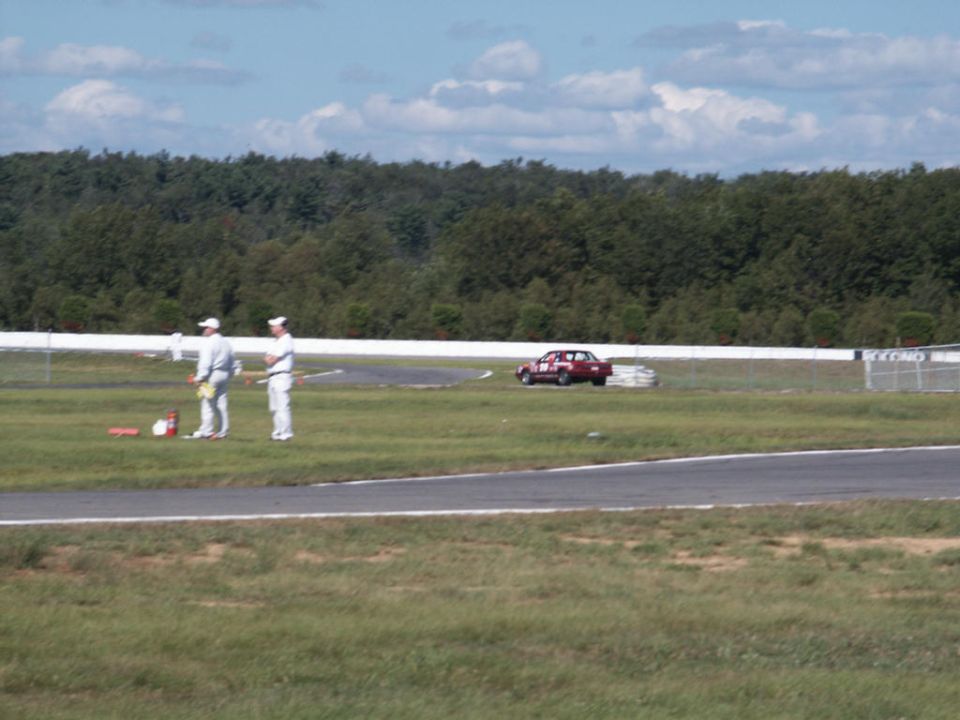 Red sedan with racing numbers on door parked on grass next to a racetrack; two people in white uniforms stand nearby