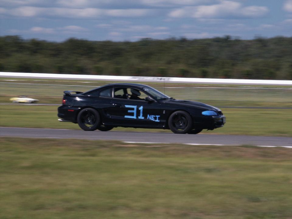 Black Ford Mustang with blue number 31 and 'NEI' on the door, on a race track. Another car is visible in the background