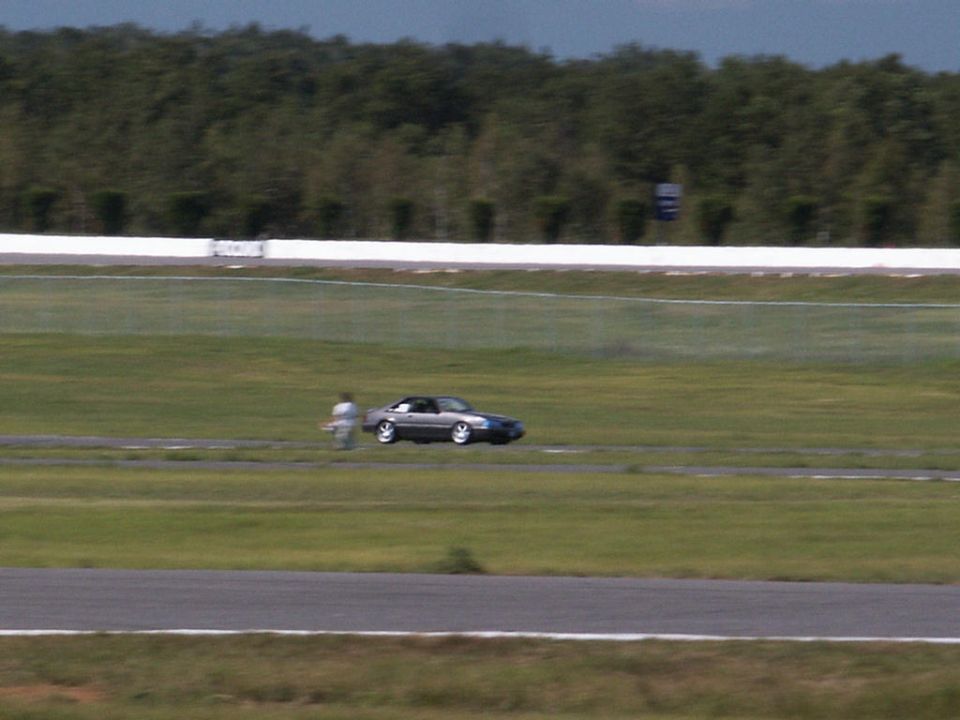 Car on a racetrack with a person nearby. Supermotors.net logo in corner