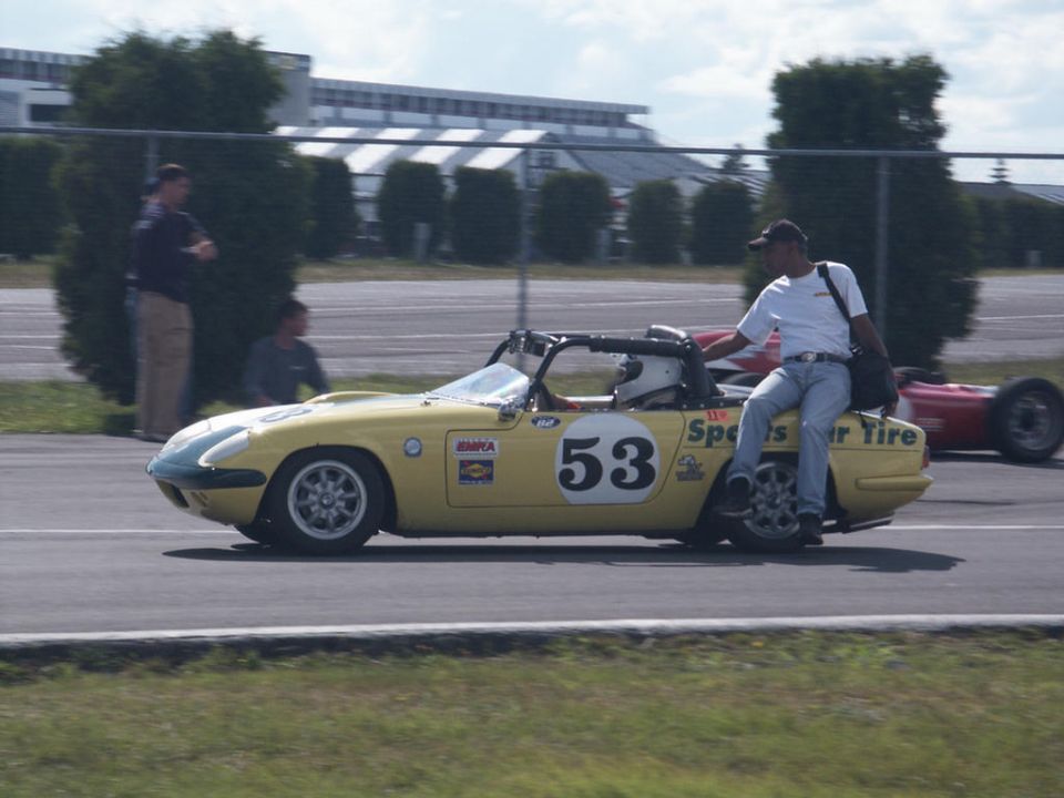 Yellow vintage sports car with racing number 53 on its side, a man sits on the rear fender. Another red race car is visible in the background