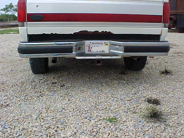Rear of white Ford with red stripe, chrome bumper, Kansas plate, and rust on tailgate, parked on gravel