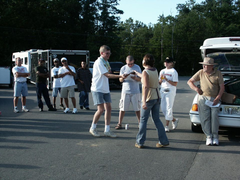 Group of people standing around in a parking lot with trailers and a car in the background