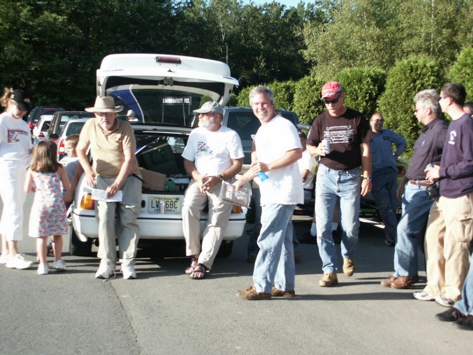 Group of people gathered around the open trunk of a white sedan. License plate reads LV 6BC
