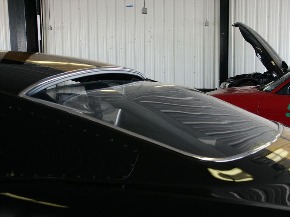 Black car's rear window and roofline with chrome trim. Red car with hood up in background