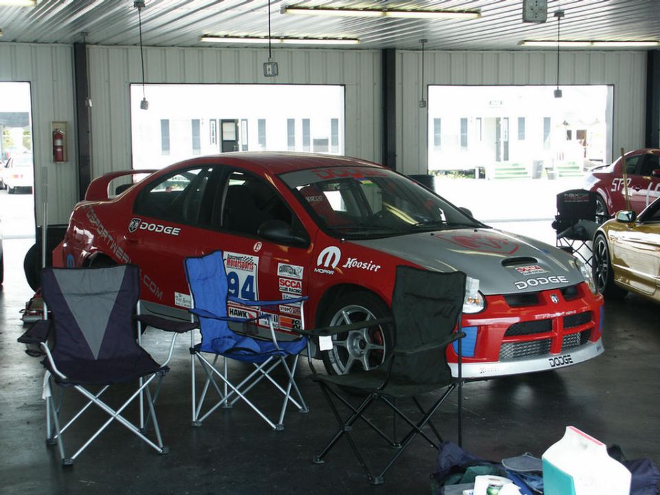 Red and silver Dodge Neon race car with number 94, parked in a garage with two folding chairs in front. Another car is visible to the right
