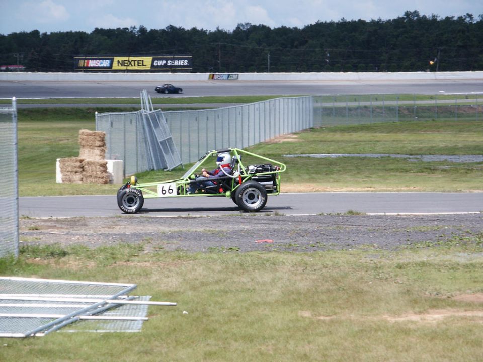 Green dune buggy with number 66 on side, driver wearing helmet, on a race track. Another car is visible in the background near NASCAR signage
