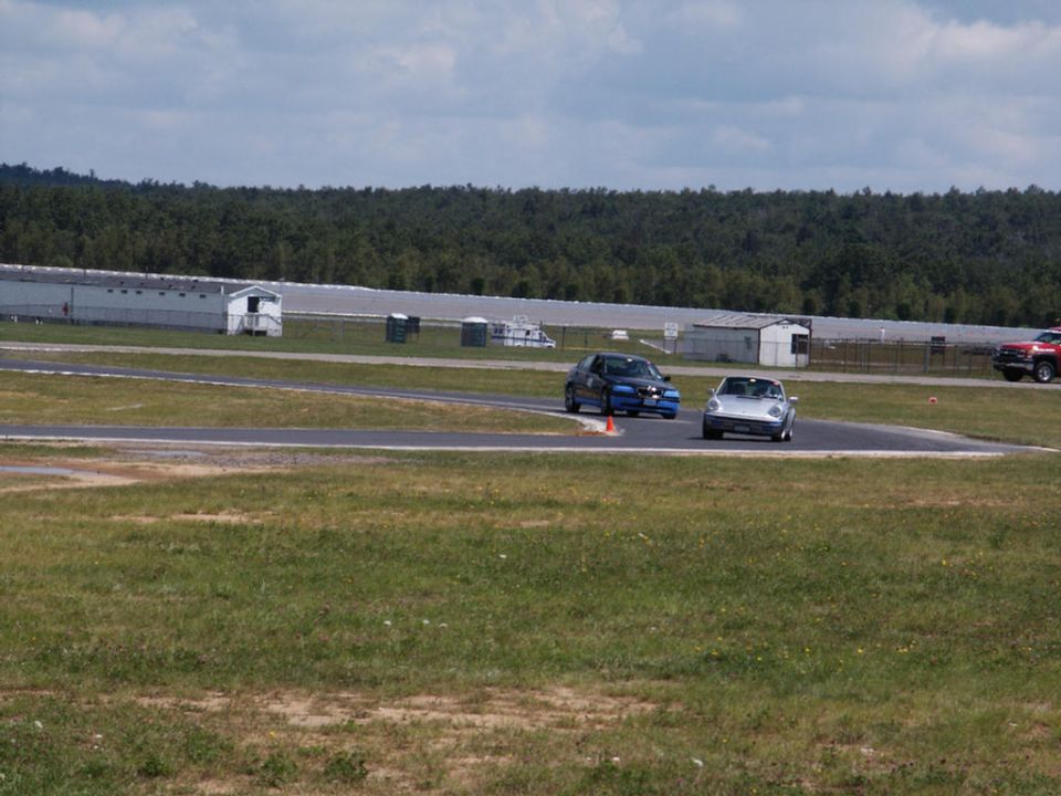 Silver convertible and blue sedan on a racetrack with buildings and trees in the background