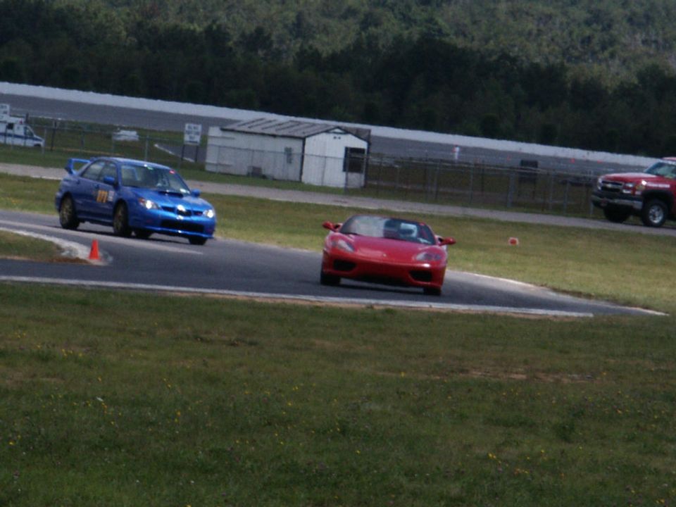 Red convertible sports car on a race track with a blue Subaru WRX and a red pickup truck in the background