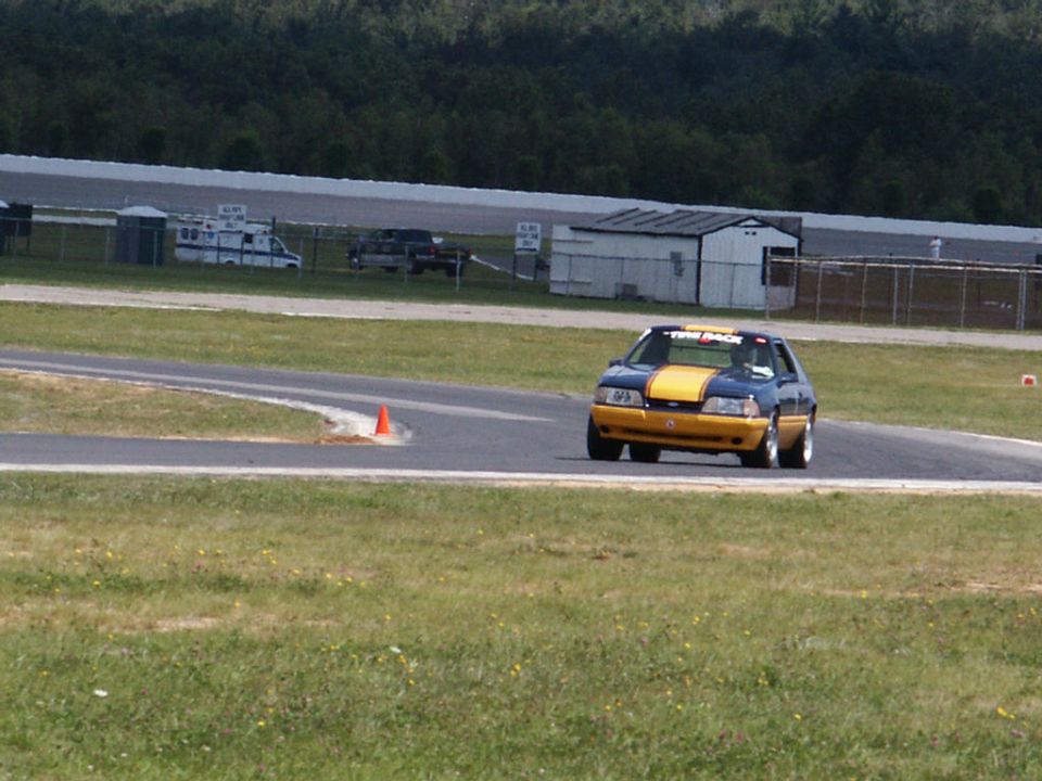Blue and yellow race car on a track, with an ambulance and truck in the background