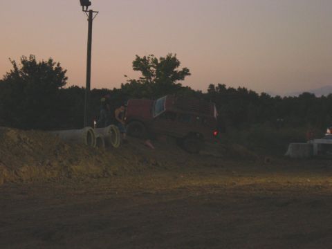 1994 Ford Bronco - A red pickup truck is positioned on a dirt mound at dusk, with two individuals standing near it.