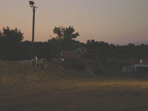 1994 Ford Bronco - A red SUV is overturned on a dirt mound at dusk, with a single person observing the scene.