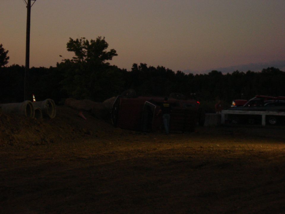Overturned red vehicle on dirt with another vehicle parked nearby at dusk