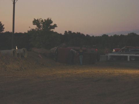 1994 Ford Bronco - A red SUV lies on its side near a flatbed truck, with a wooded area in the background.