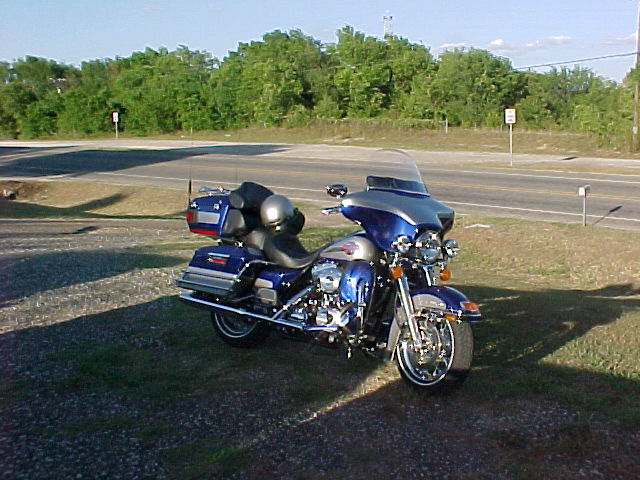 Blue and silver touring motorcycle parked on grass, with saddlebags and a helmet on the seat. Road and trees in background